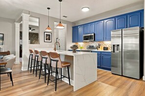 A modern kitchen with blue cabinets, a stainless steel refrigerator, and a marble island with bar stools. - Luxury 3BR Modern Design Lake Views Vista Cay (Orlando)