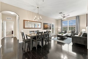 A spacious living room with a dark wood floor, a dining area with a wooden table and chairs, a ceiling fan, and a large window with curtains. - 3BR Vista Cay Conventions Parks and Orlando Fun (Orlando)