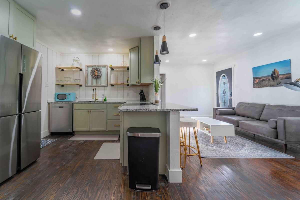Una cocina moderna con una isla central, electrodomésticos de acero inoxidable y un sofá gris en la sala de estar.
