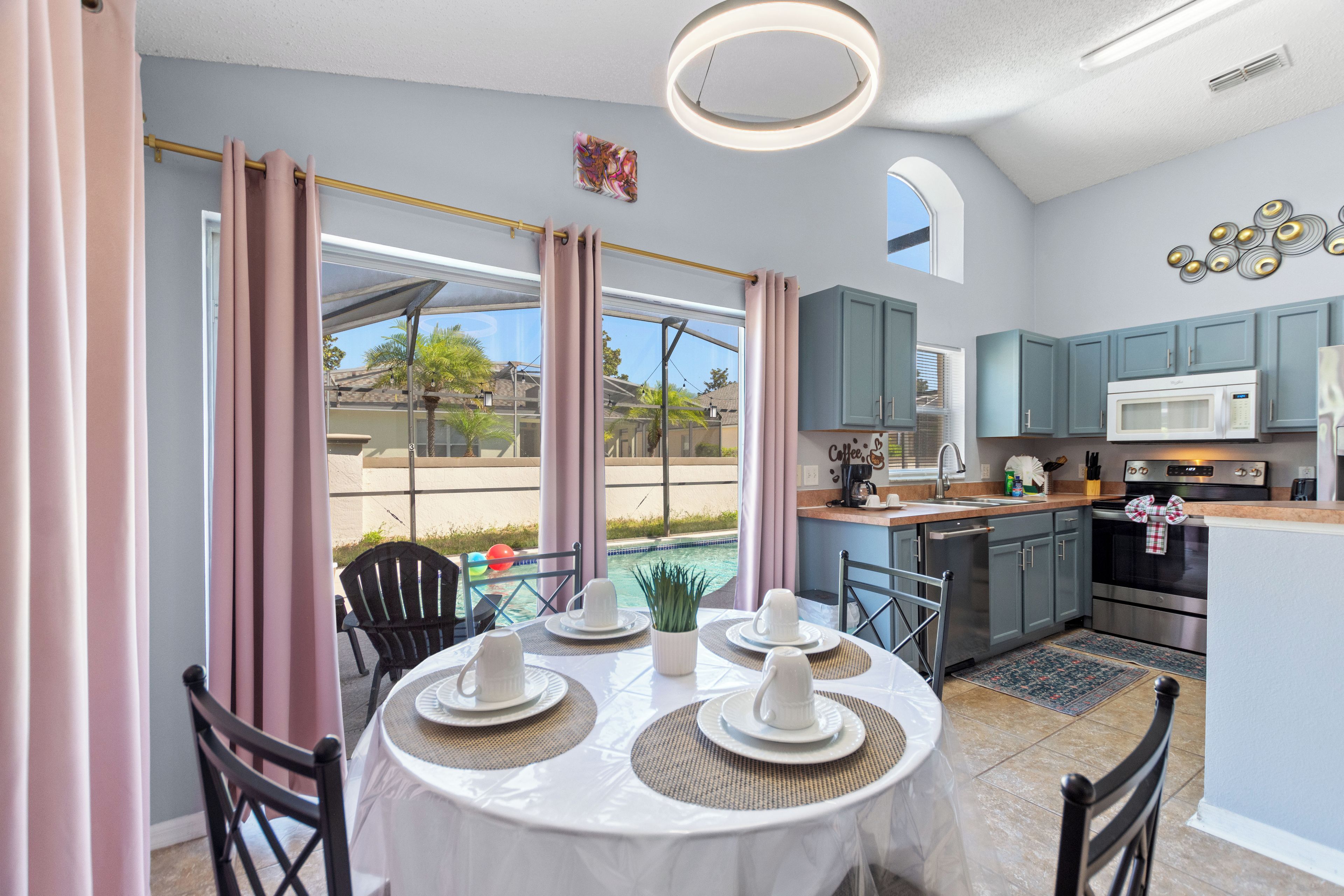 A modern kitchen with a dining area, featuring a round table with white tablecloths and plates, a large window with curtains, and a view of the outdoors.