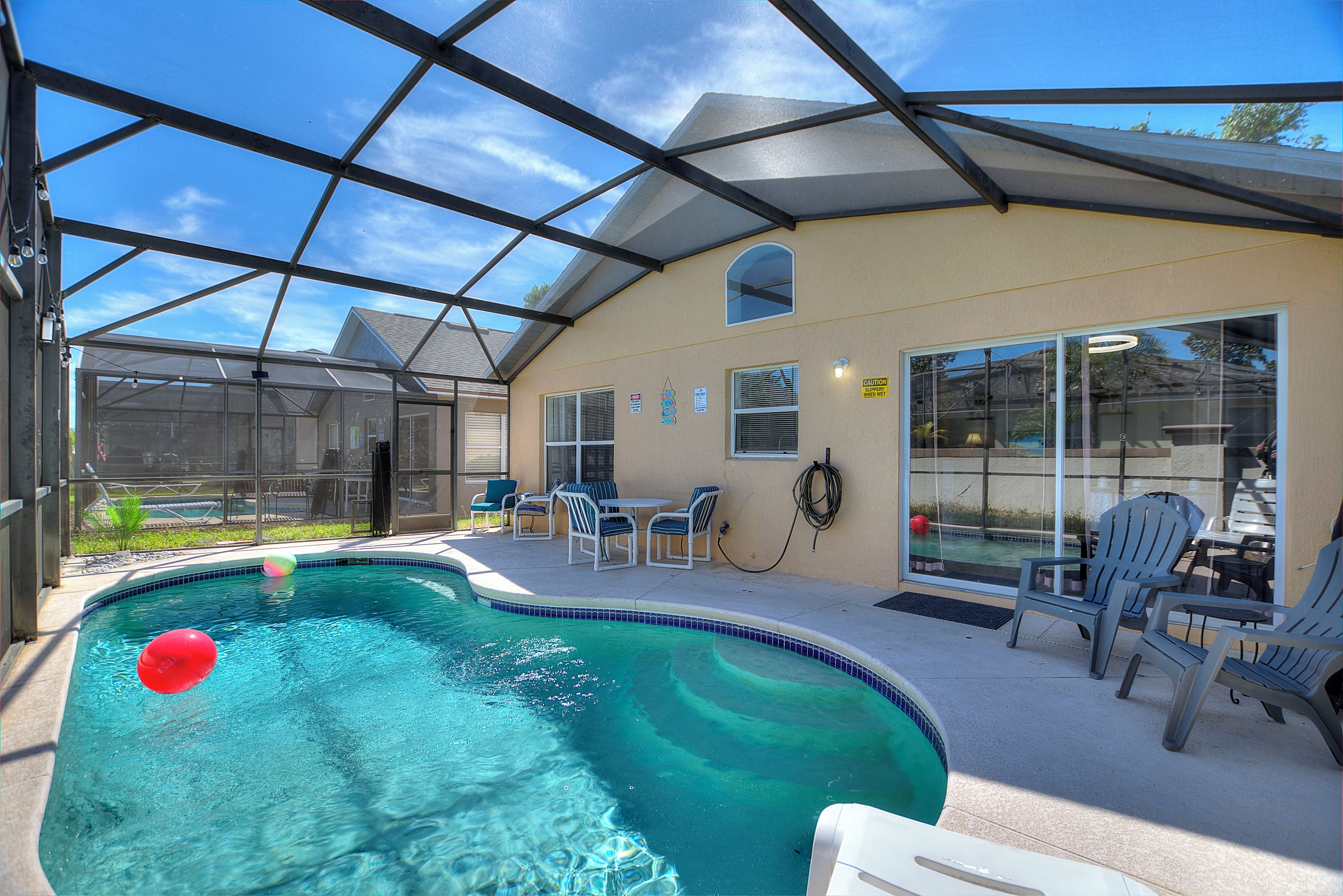 A covered swimming pool with a glass enclosure, adjacent seating area, and a house in the background.