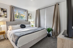 Quarto de hotel moderno com uma cama grande, uma mesinha de cabeceira com abajur, uma planta em vaso e uma TV sobre um suporte de madeira.