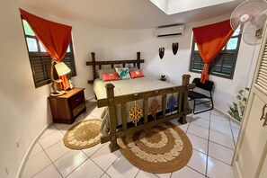 Una camera da letto con un letto, un comodino, una lampada, cuscini decorativi, una testiera in legno, una ventola e una finestra con tende.