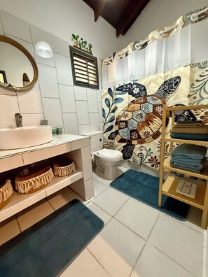 Un bagno con lavandino, specchio e water bianchi. Si nota uno scaffale con asciugamani piegati e un tappetino da bagno blu sul pavimento.
