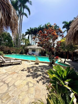 Un'area piscina con lettini, una struttura con tetto di paglia e vegetazione tropicale.