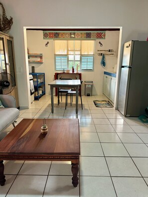 Una sala de estar con una mesa de centro de madera, un sofá, una mesa de comedor con sillas, una zona de cocina con un refrigerador y una ventana con persianas.