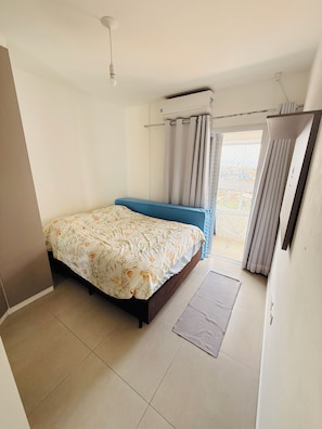A bedroom with a bed, a window with curtains, an air conditioning unit, and a view of the outside. - Ocean View | Cozy and Steps from the Beach (Praia Grande)