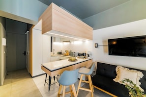 A modern kitchen with a built-in oven, a sink, and a countertop with a coffee maker. There is a dining area with a table and two chairs. A television is mounted on the wall. - Large Terrace Studio · Selected Design (H03) (Providencia)
