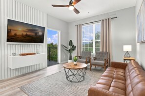 Ein modernes Wohnzimmer mit einer Ledersofa, einem runden Couchtisch, einem an der Wand montierten Fernseher und einem Balkon mit Vorhängen.