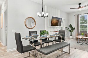 Ein moderner Essbereich mit einem Glastisch, schwarzen Stühlen, einem spiegelnden Wandbild mit Holzrahmen und einem Deckenventilator.