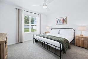Un dormitorio con cama, mesita de noche, lámpara, ventana con persianas, ventilador de techo y un cuadro colgado en la pared.