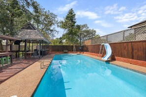 A rectangular swimming pool with a slide, surrounded by a wooden deck and a covered seating area. - Aggieland Oasis: Sleeps 12, Pool, Hot Tub, 7 Min to Texas A&M (College Station)