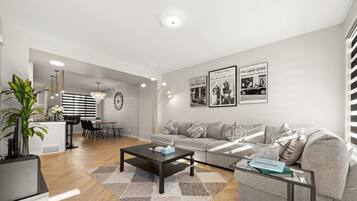 A modern living room with a sofa, coffee table, and dining area. There are wall-mounted lights and framed pictures.