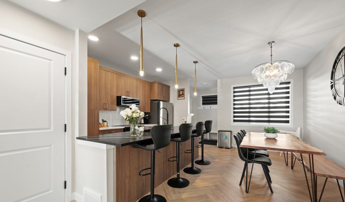 A modern kitchen with wooden cabinets, a black countertop, and bar stools. There is a dining table with chairs and a chandelier above.