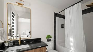 A modern bathroom with a black marble sink, a gold-framed mirror, a small potted plant, and a shower curtain.