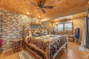 A bedroom with a stone wall, wooden ceiling, a bed with a patterned bedspread, a wooden nightstand, a dresser, and a mannequin. - Honeymoon Cabin w/MTN Views*Hot Tub*Games*Peaceful (Sevierville)