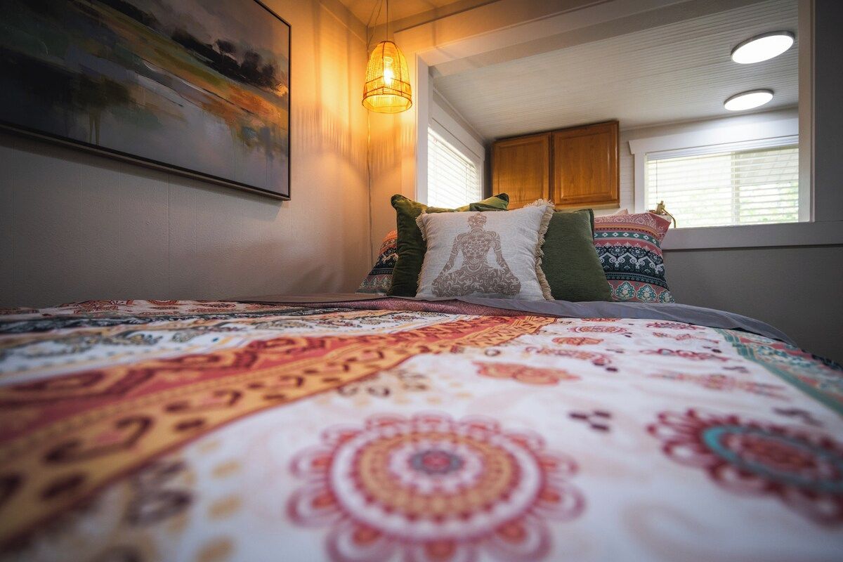 A bedroom with a patterned bedspread, a hanging light fixture, and a window with blinds.