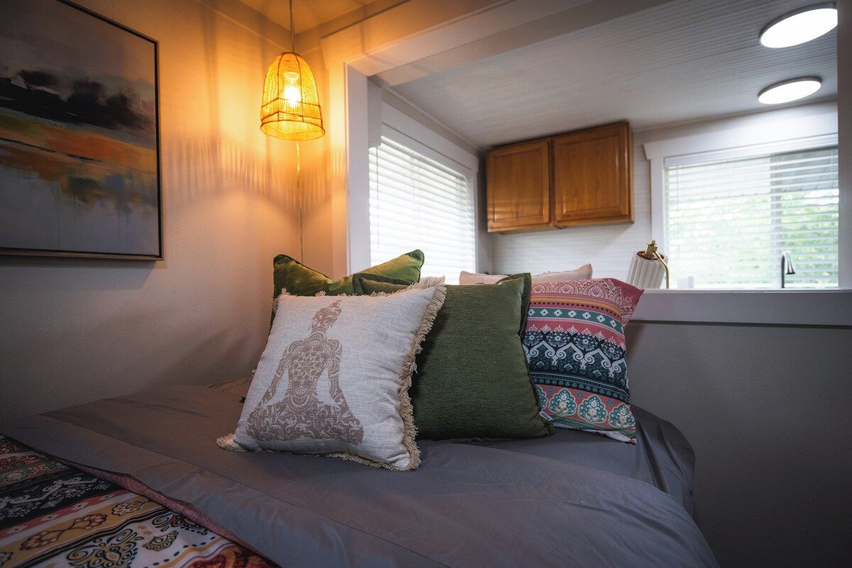 A bedroom with a bed, pillows, a wall-mounted lamp, a window with blinds, and a painting on the wall.