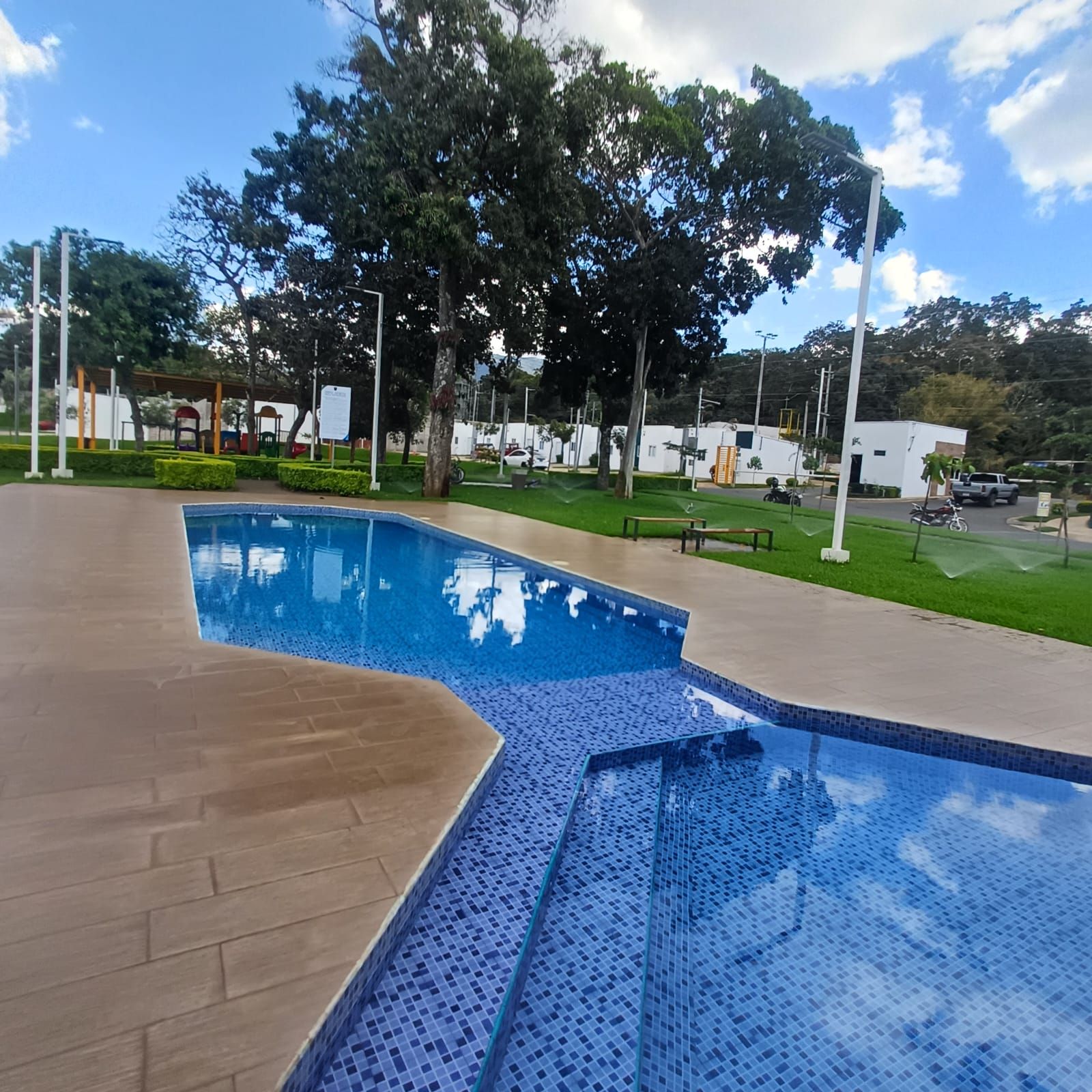 Una piscina con borde de azulejos, rodeada por un muelle de madera y una zona verde con árboles.