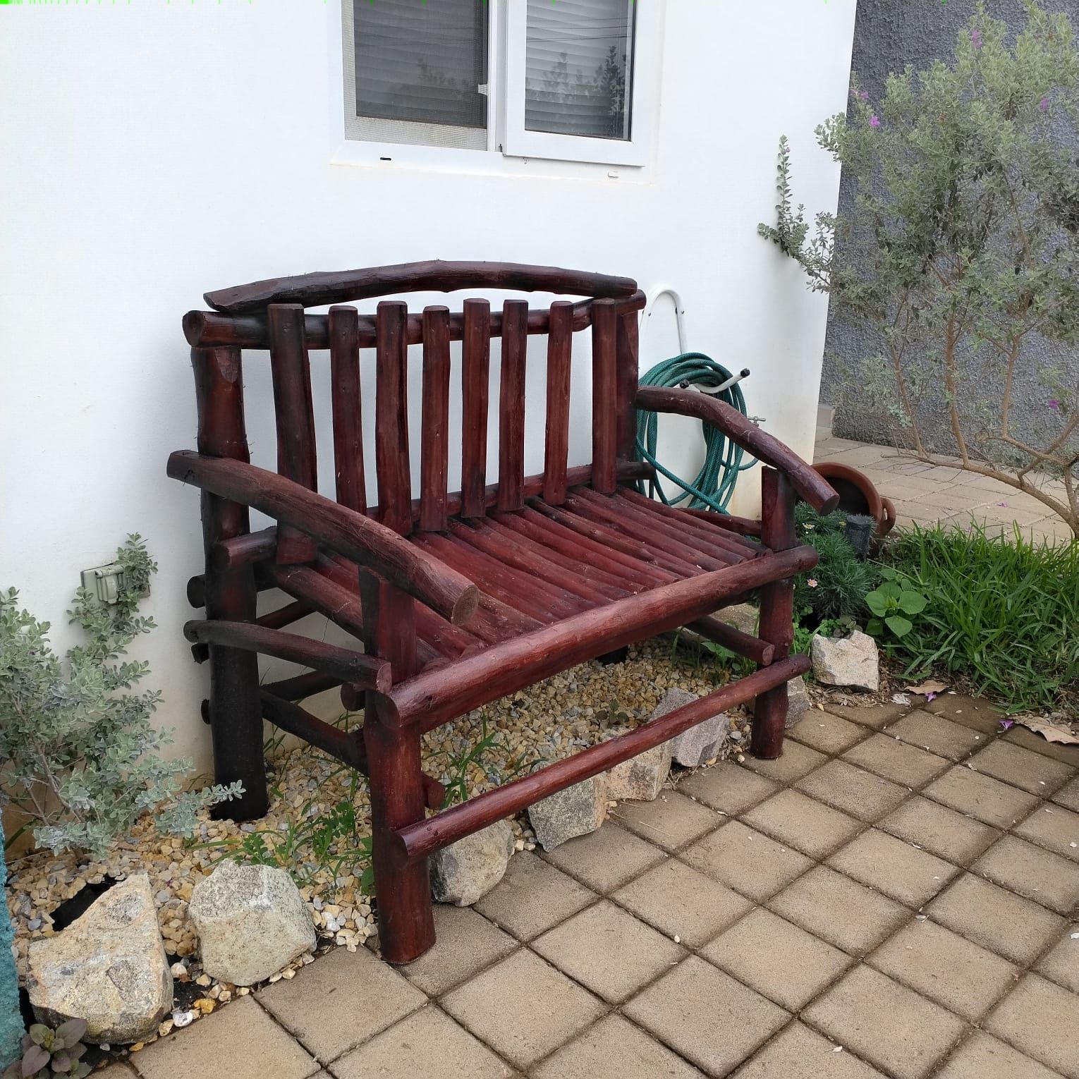 Un banco de madera con respaldo y apoyabrazos, ubicado en una zona empavedada con una pared blanca y una ventana al fondo.