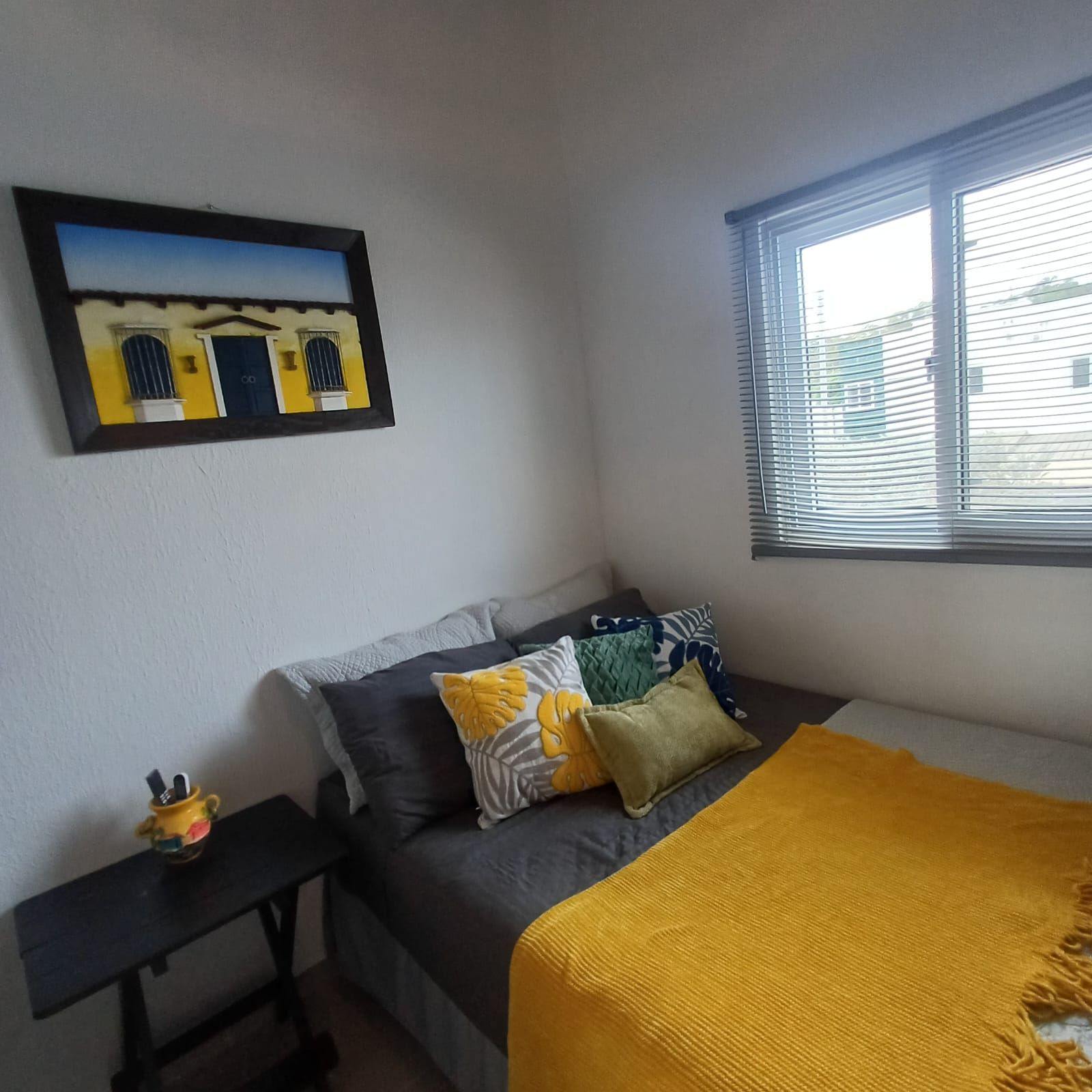 Une chambre à coucher avec un lit, des oreillers, une table de chevet, une fenêtre avec des stores et un tableau accroché au mur.