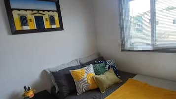 Une chambre à coucher avec un lit, des oreillers, une table de chevet, une fenêtre avec des stores et un tableau accroché au mur.