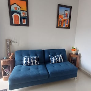 Un sofá azul con dos cojines estampados, una pequeña mesita con una planta y dos cuadros en la pared.