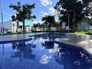 Una piscina con borde de azulejos, rodeada de vegetación y edificios.