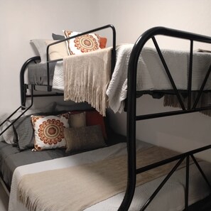 Una cama litera con una colcha blanca y una manta color beige. Hay almohadas con estampado floral y una lisa.