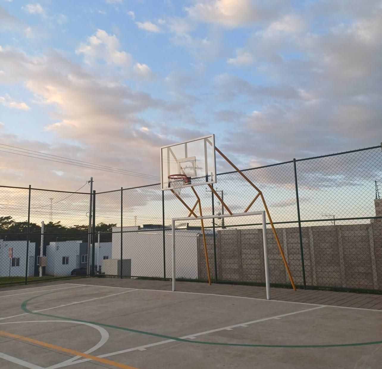 An outdoor basketball court with a hoop, surrounded by a chain-link fence, under a cloudy sky.