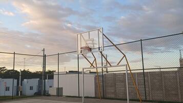 An outdoor basketball court with a hoop, surrounded by a chain-link fence, under a cloudy sky.