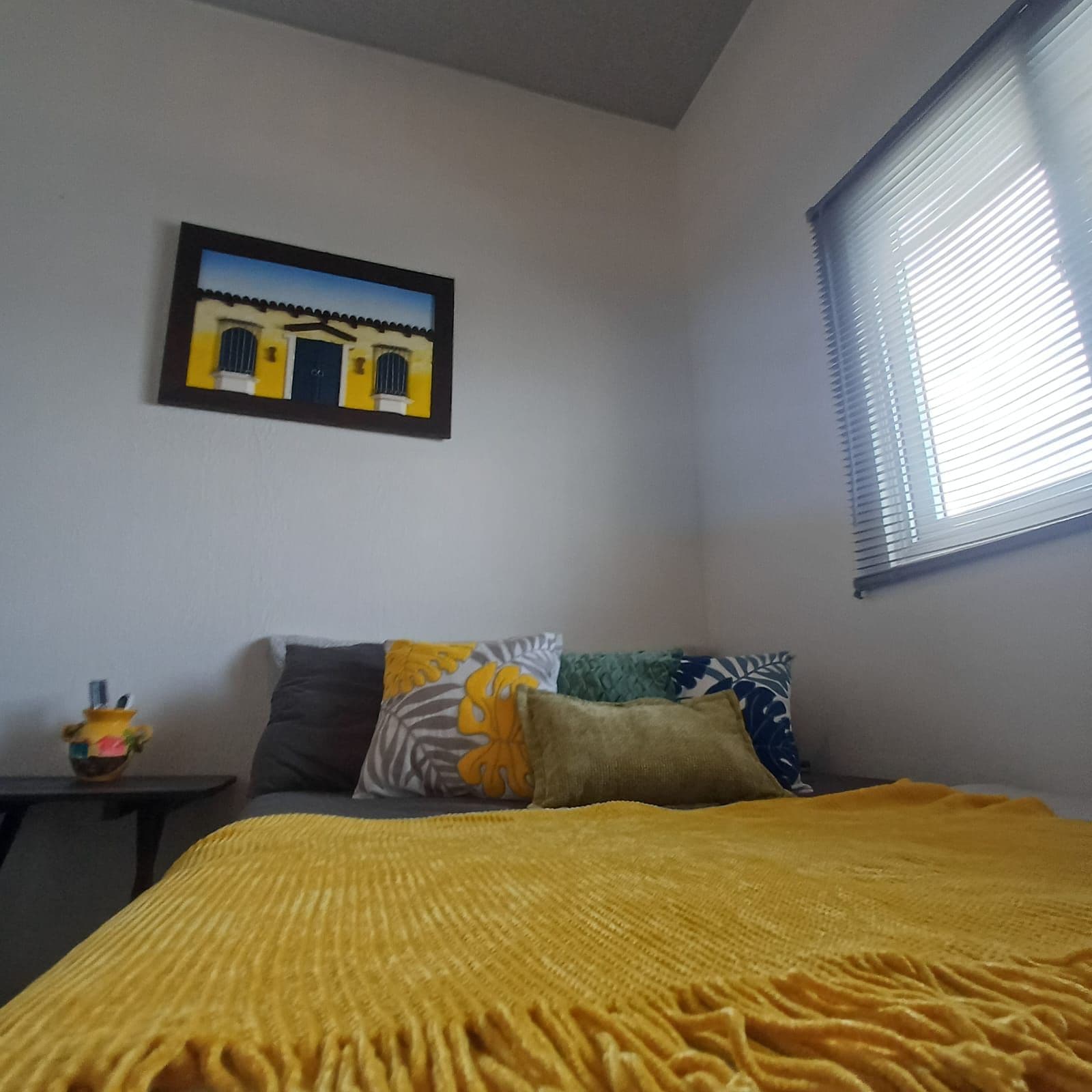 A bedroom with a bed, pillows, a small table with a decorative item, a window with blinds, and a wall-mounted picture.