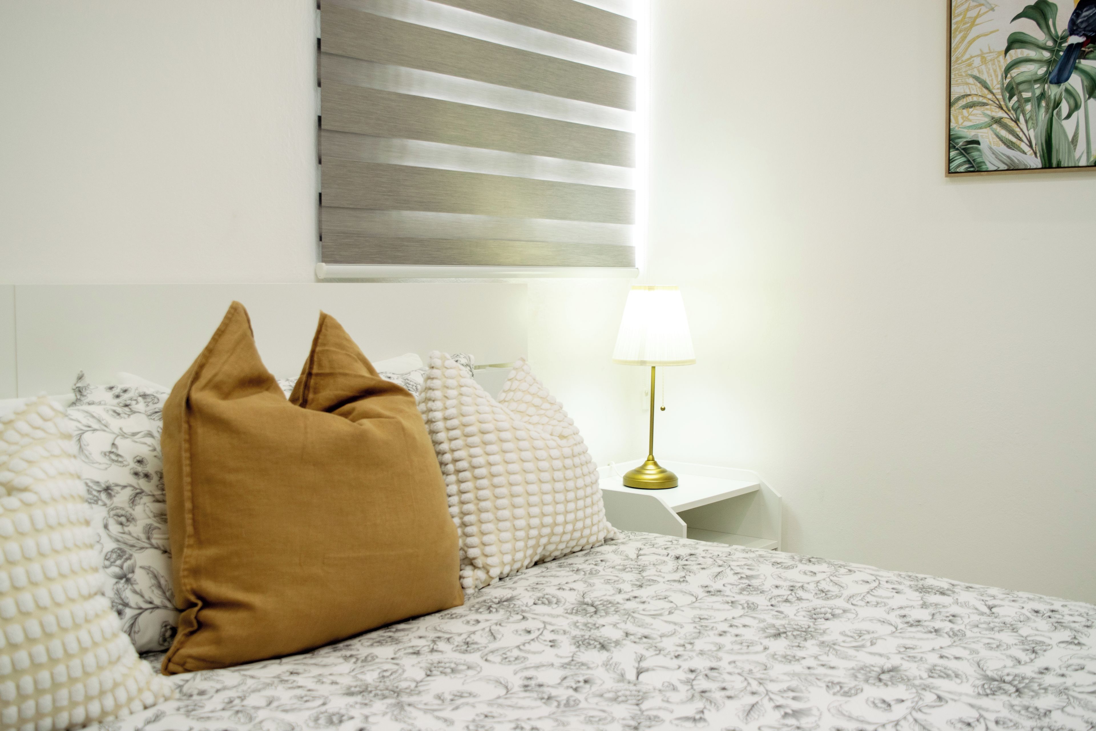 Cama com roupa de cama estampada, um único travesseiro marrom, abajur e janela com persianas.