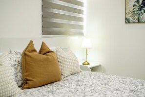 Ein Bett mit gemusterten Bettbezügen, ein einzelnes braunes Kopfkissen, eine Nachttischlampe und ein Fenster mit Jalousien, das an der Wand montiert ist.