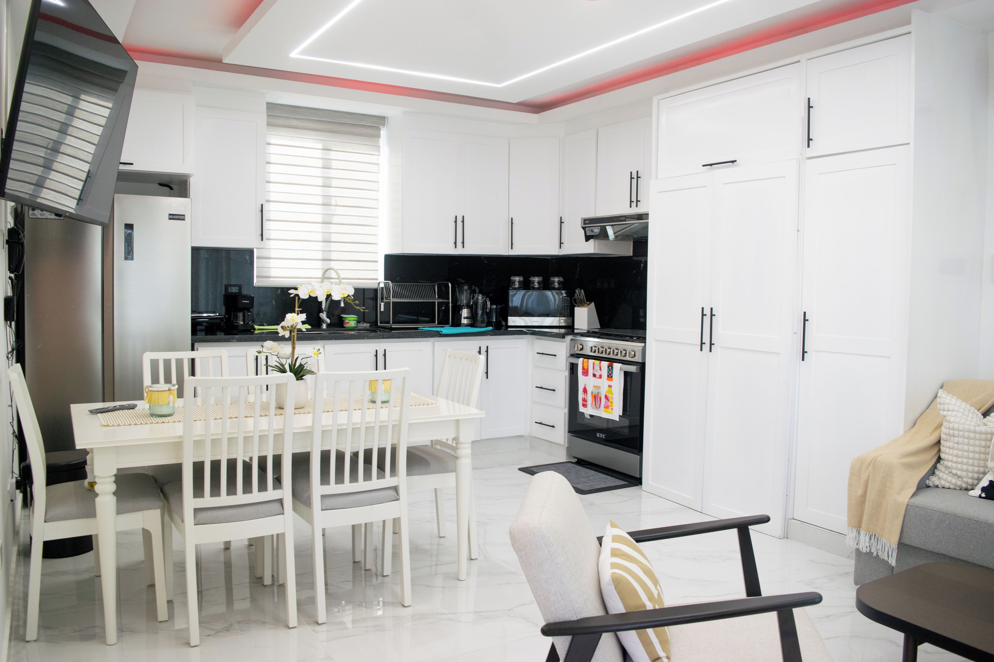 Una cocina moderna con armarios blancos, una mesa de comedor con sillas, horno empotrado y cafetera.