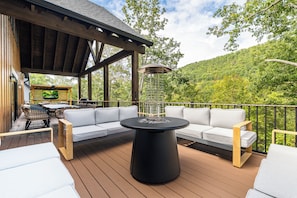 A deck with a sofa and a round table, overlooking a forested hillside.