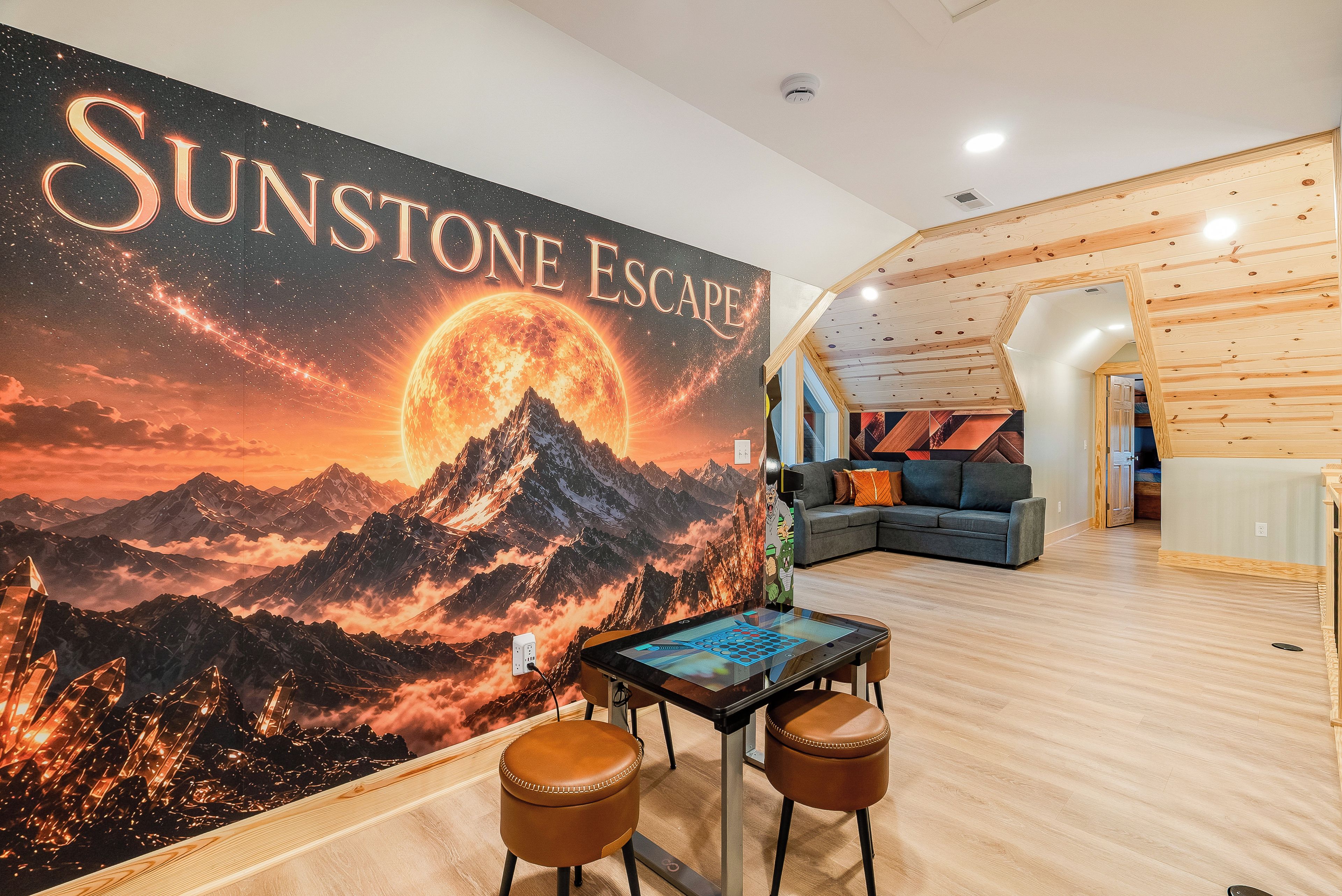 A room with a large wall mural titled 'Sunstone Escape,' a wooden ceiling, a sofa, and a table with stools.