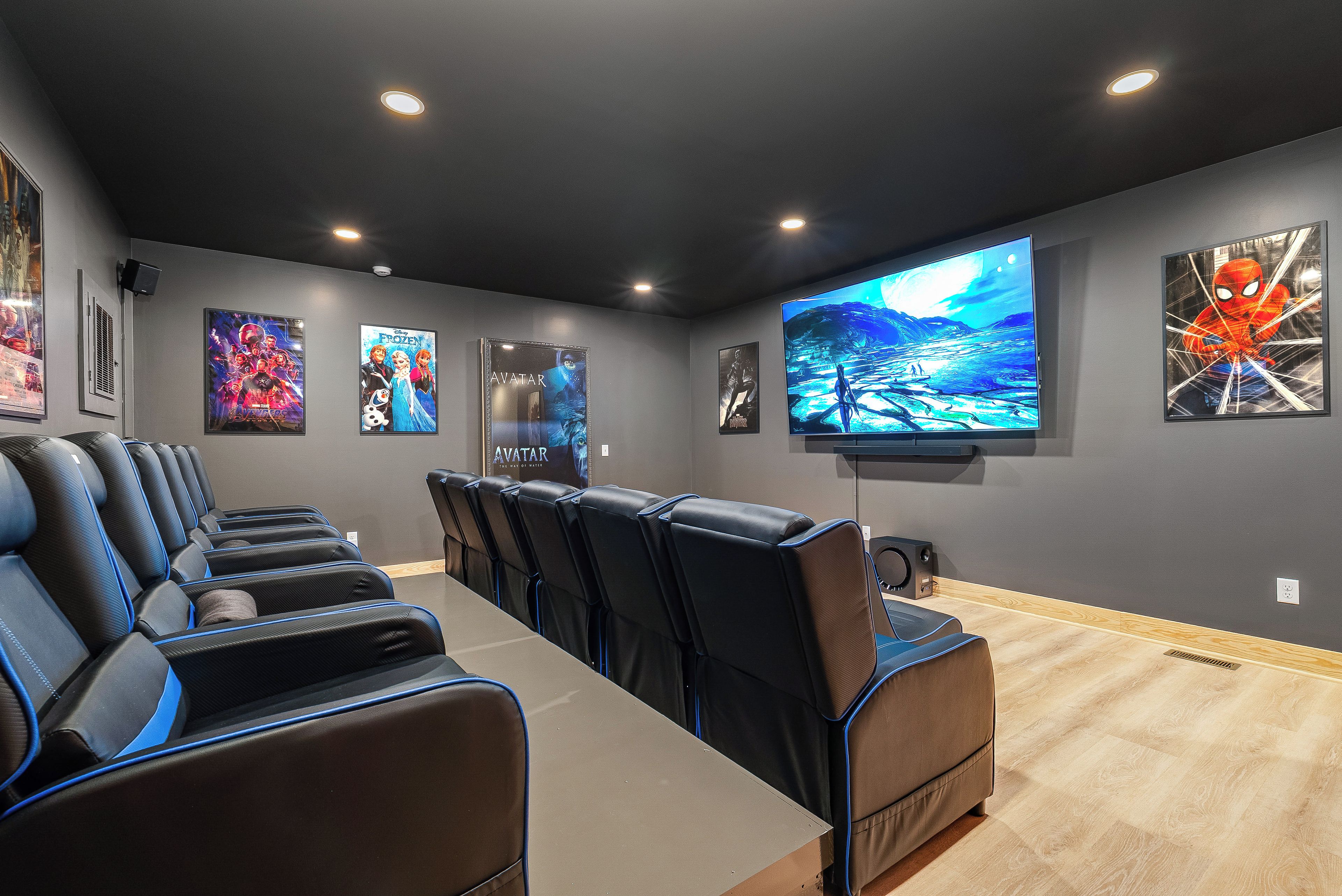 A home theater room with a large flat-screen TV, multiple posters on the wall, and a row of black leather recliner chairs.