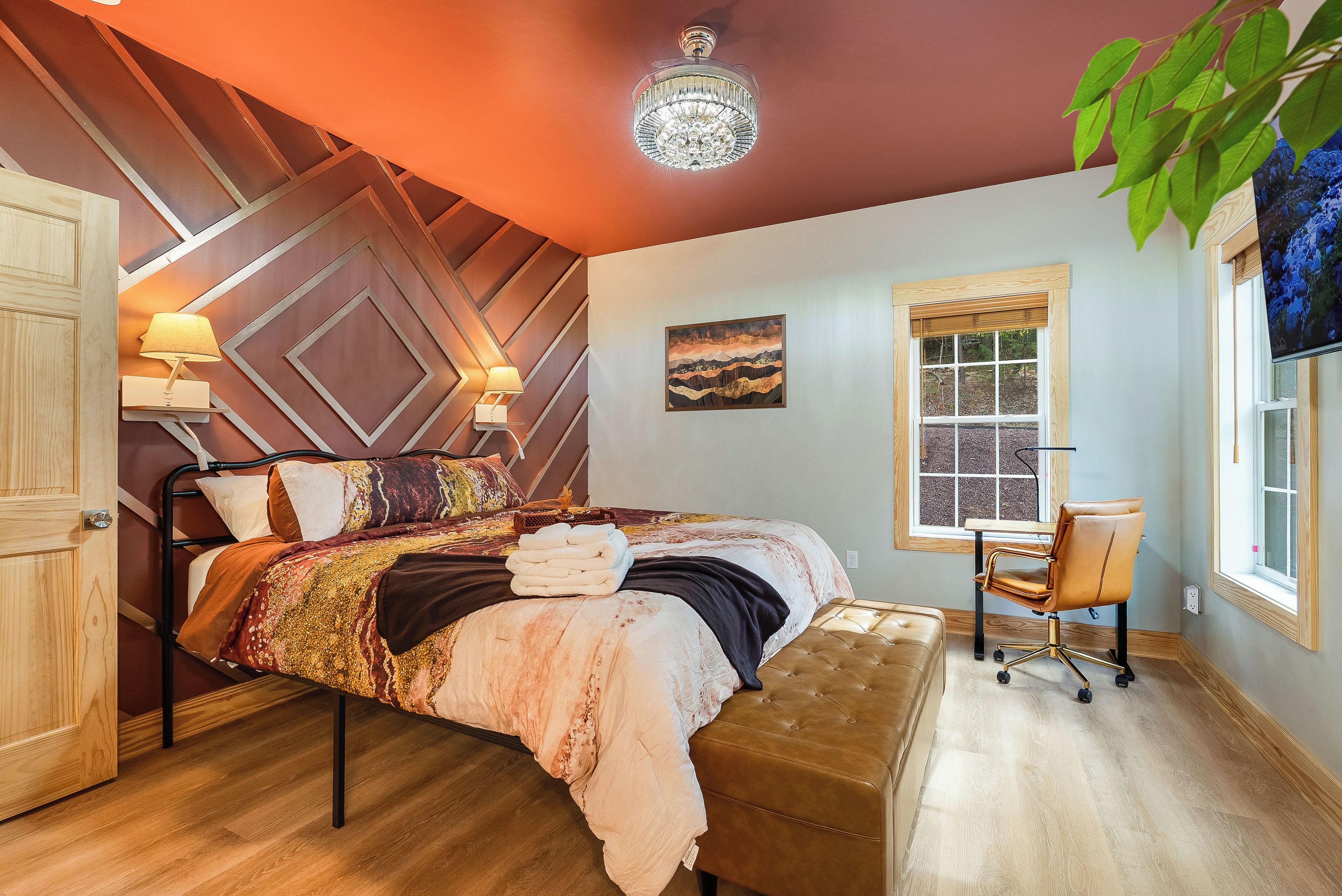 A bedroom with a bed, bedside lamps, a desk, a chair, a window, and a chandelier.