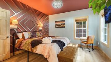 A bedroom with a bed, bedside lamps, a desk, a chair, a window, and a chandelier.