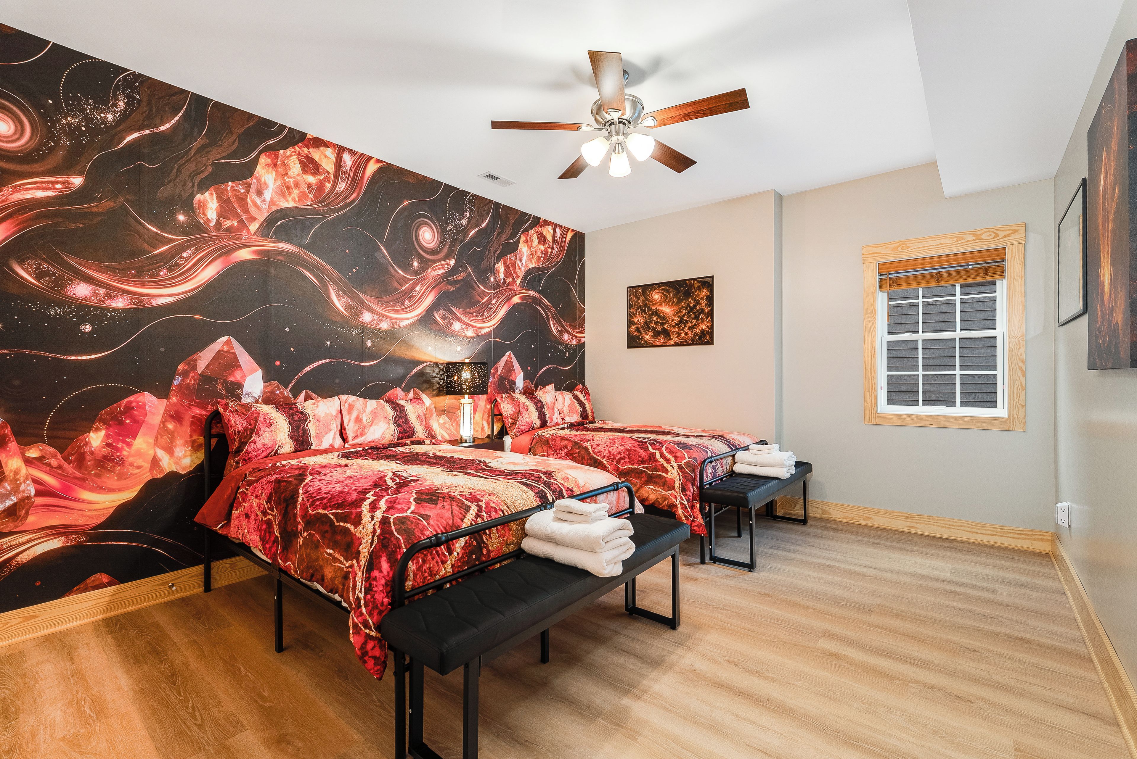 A room with two beds, a ceiling fan, a window with blinds, and a wall with a large abstract painting.