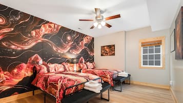A room with two beds, a ceiling fan, a window with blinds, and a wall with a large abstract painting.