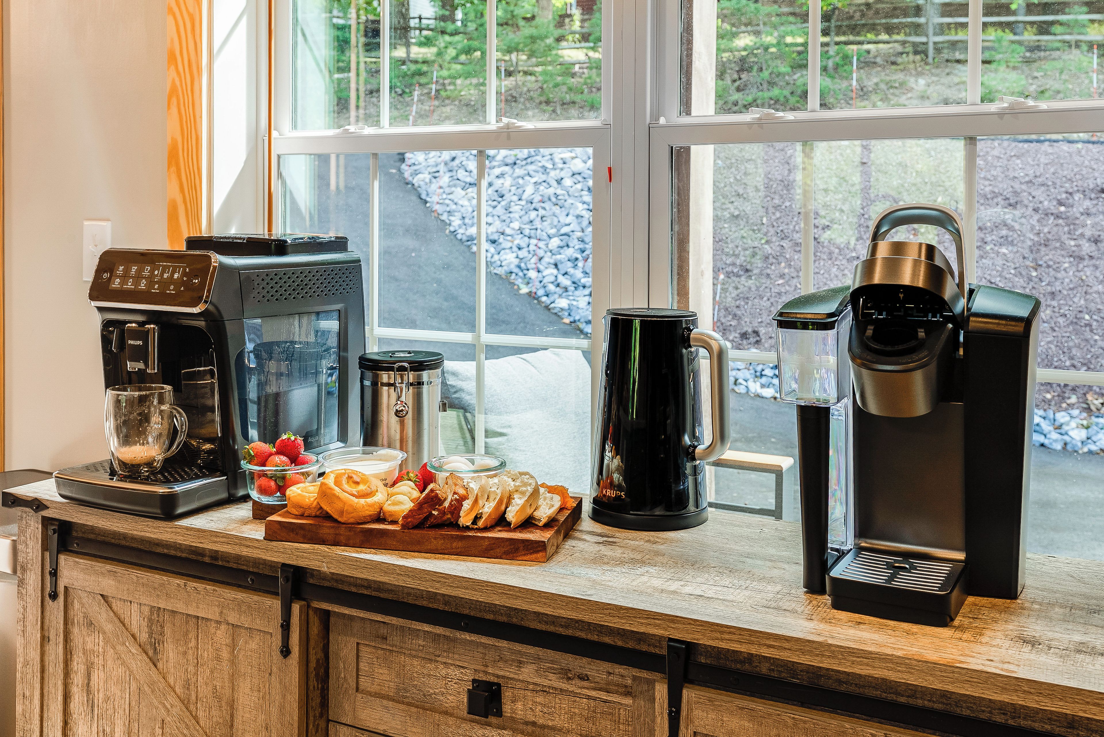 Kaffestation med kaffemaskine, mælkeskummer, kaffekande og et fad med kager og frugt.
