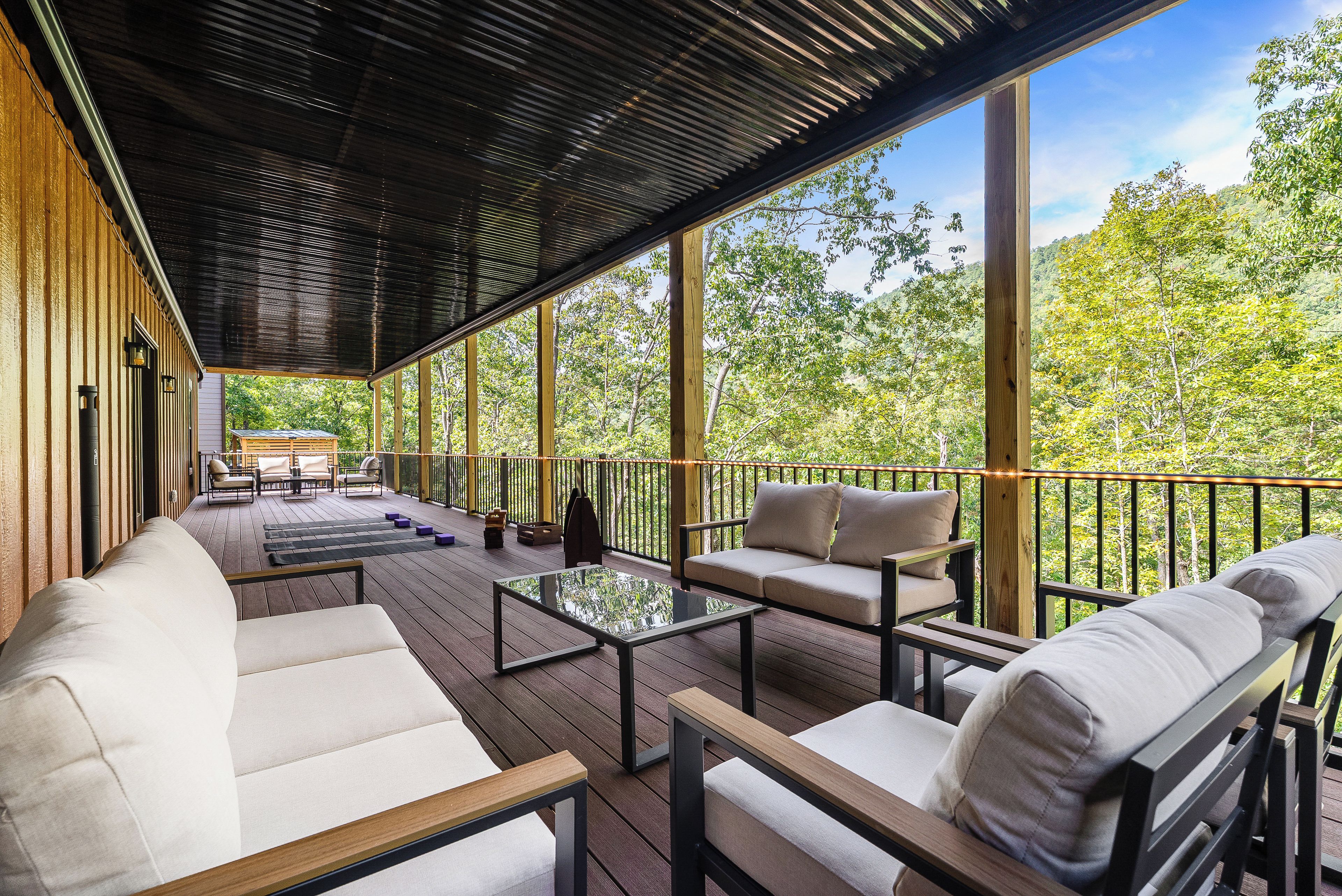 A covered outdoor seating area with a sofa, chairs, and a table, overlooking a forested landscape.