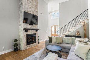 A modern living room with a stone fireplace, a flat-screen TV, a staircase, and a sofa with cushions. - Spacious 5BR Near Liberty University, PS5 & PatioTable Fire Pit, Pack-n-Play (Lynchburg)