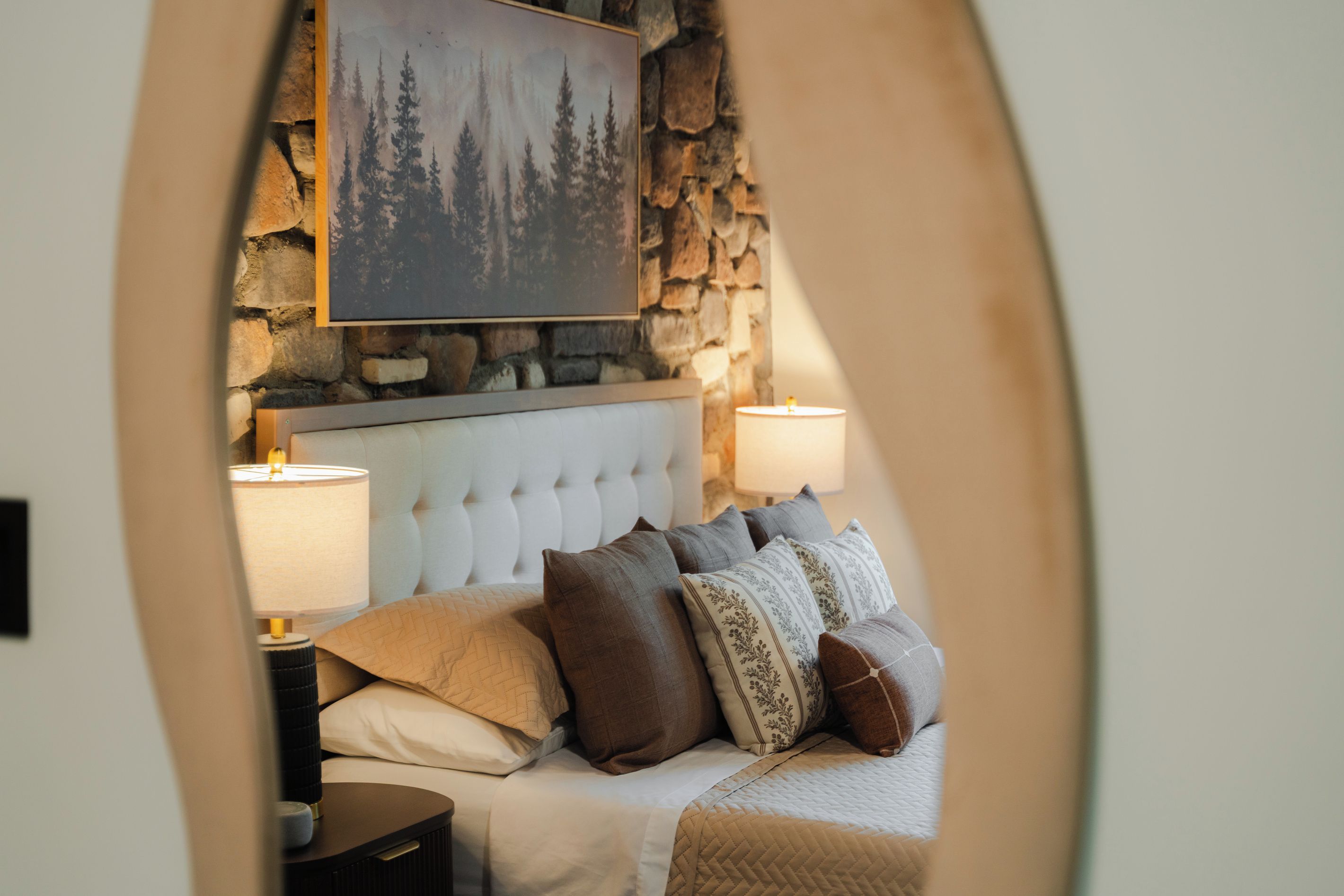 Ein Schlafzimmer mit einer Steinwand, einem rautenförmig gepolsterten Kopfteil und einem Gemälde eines Waldes. Es gibt zwei Nachttischlampen und mehrere Kissen.