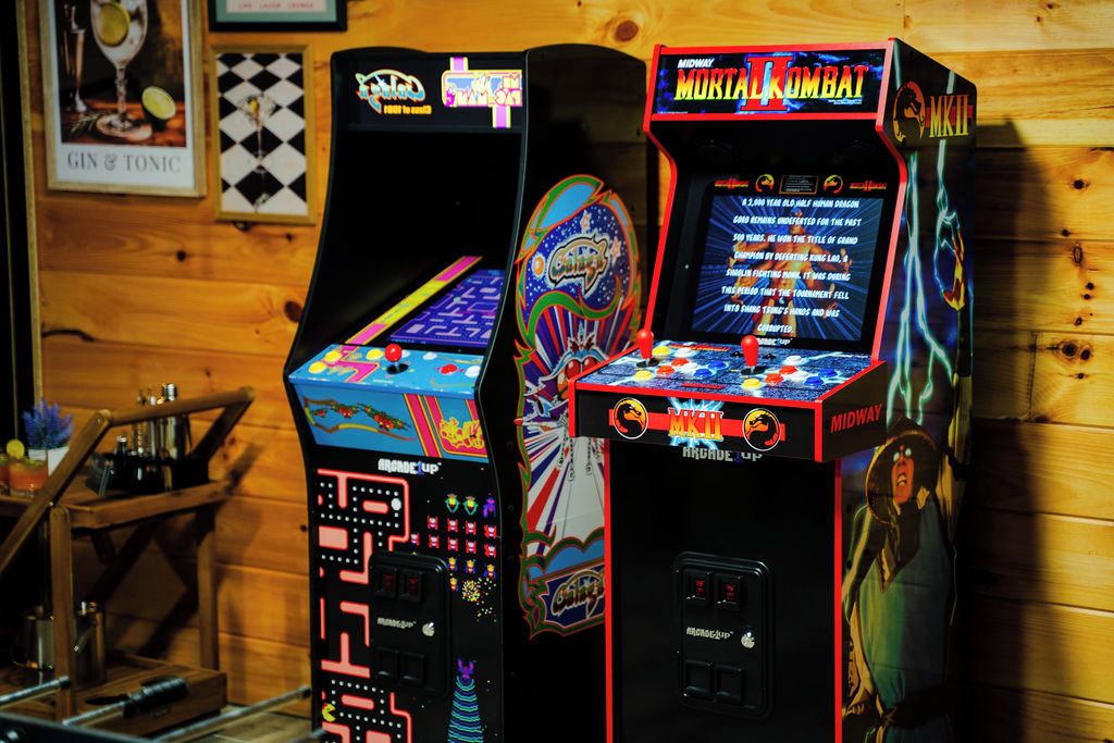 Zwei Spielautomaten, einer mit der Aufschrift 'Midway' und der andere mit 'Mortal Kombat', vor einer Holzwand.