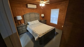 A bedroom with a wooden headboard, a bed with a white bedspread, two bedside lamps, a nightstand, a ceiling fan, and an air conditioning unit. - Base Camp on Cherry Hill — Fife Lake, MI 

Cabin, Sleeps 4–8 (Fife Lake)
