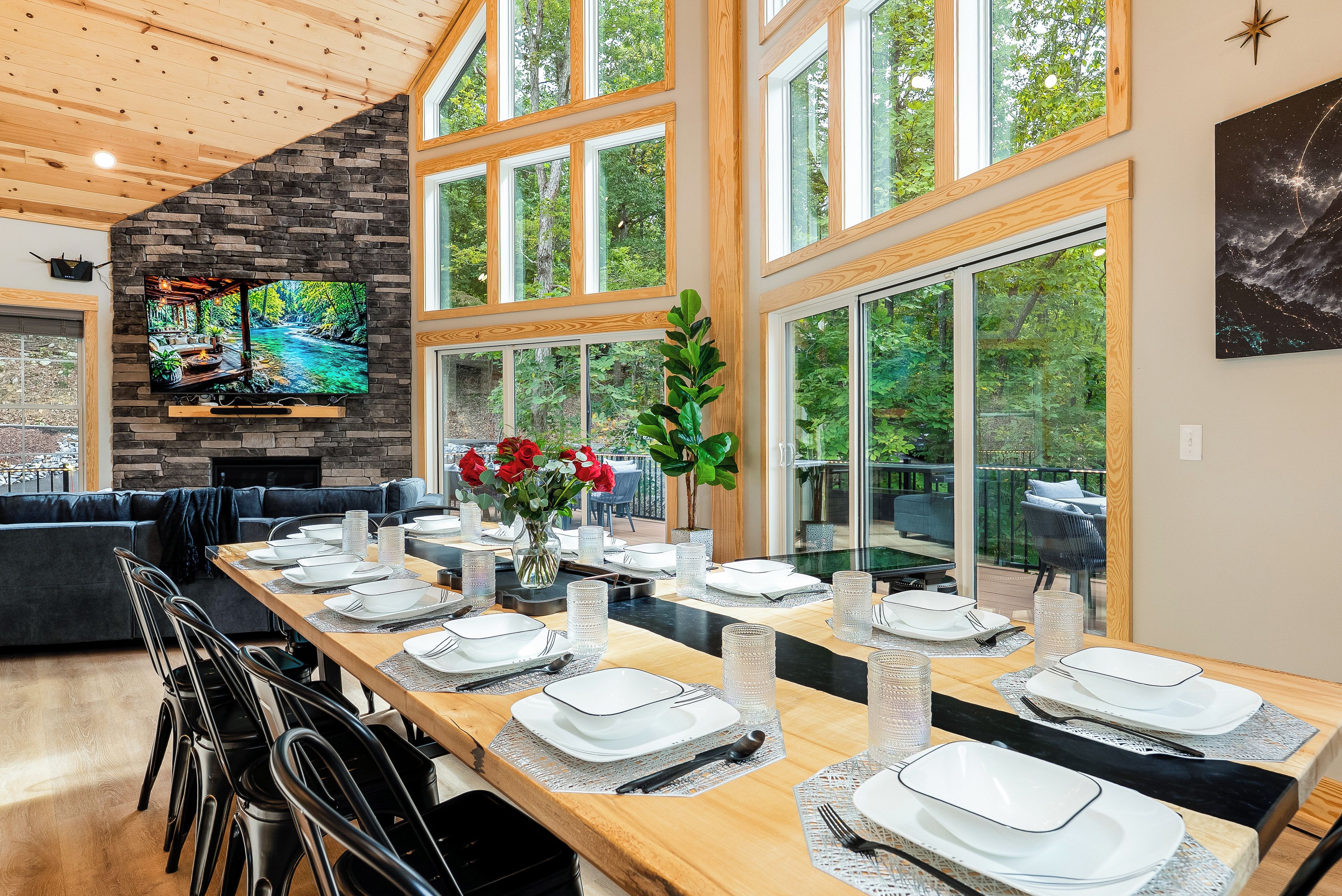 A dining area with a long wooden table set for a meal, a stone fireplace, and large windows offering a view of the outdoors.