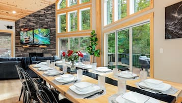A dining area with a long wooden table set for a meal, a stone fireplace, and large windows offering a view of the outdoors.